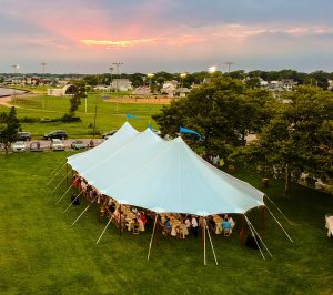 Sailcloth marquee set up on a grassy field with wooden poles, photographed from above during an outdoor evening event.